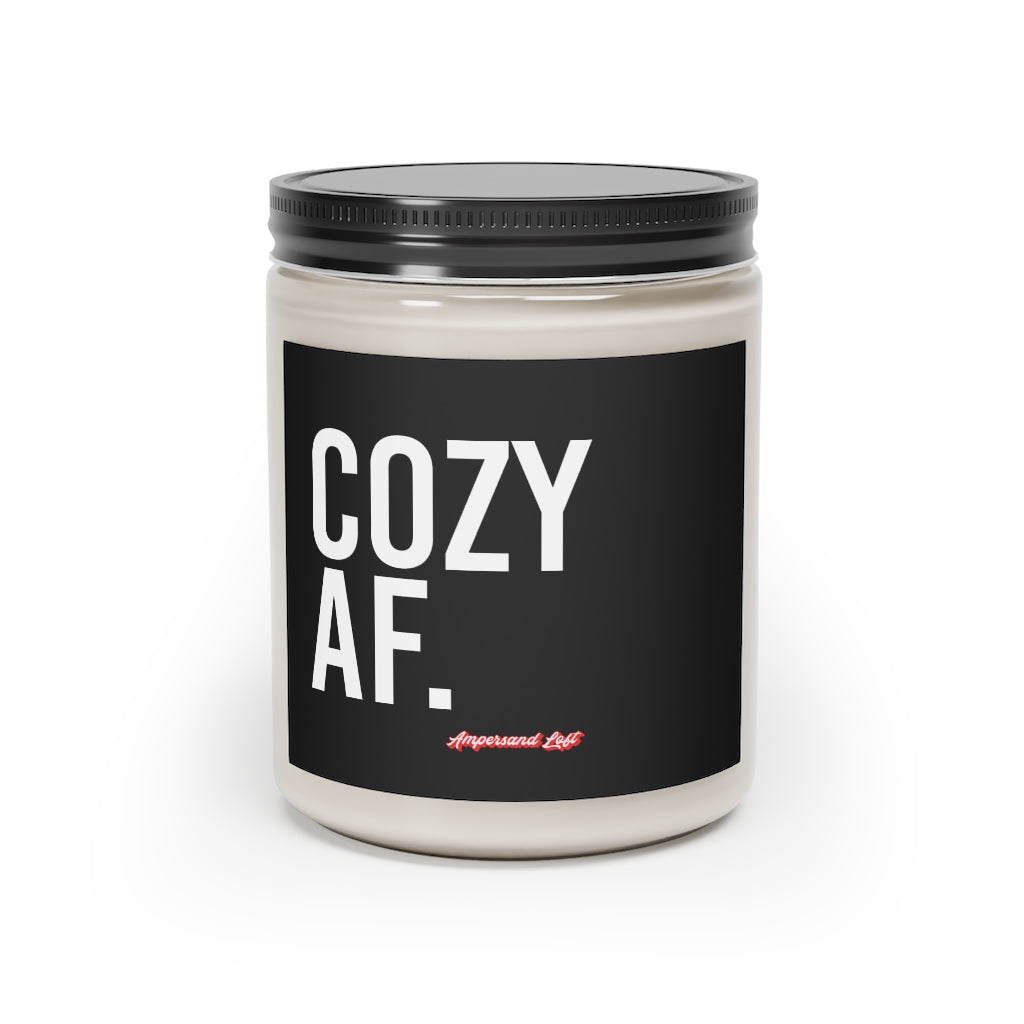 Candle in a glass jar with black label that reads, in white All-Caps text "Cozy AF." and a small "Ampersand Loft logo below (pink retro font with solid red drop shadow)
