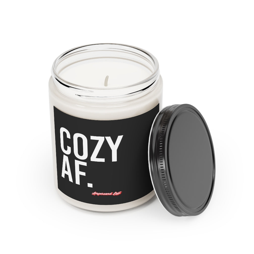 Candle in a glass jar with the lid off, resting on the side of the candle. Black label that reads, in white All-Caps text "Cozy AF." and a small "Ampersand Loft logo below (pink retro font with solid red drop shadow)