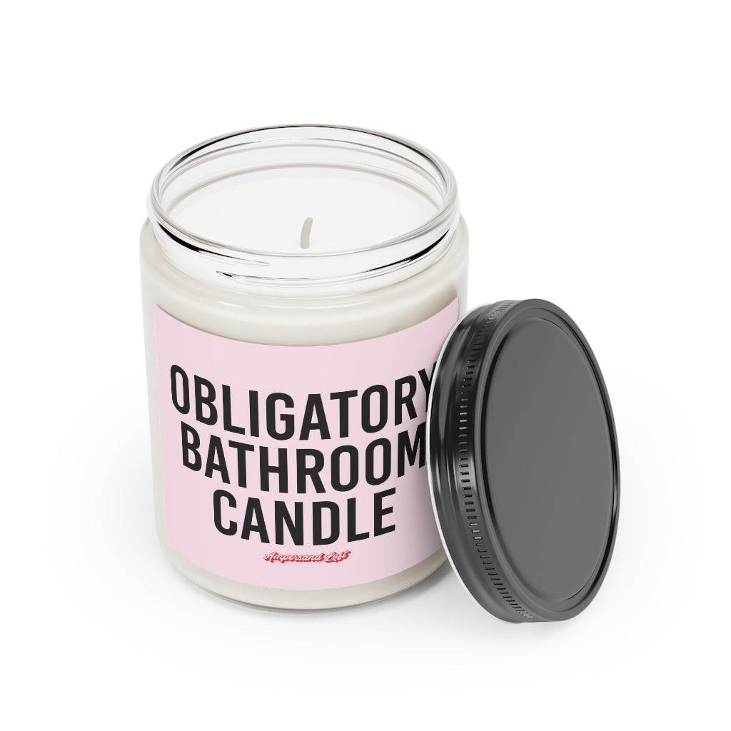 Candle in a glass jar with the lid off, resting on the side of the candle. Pink label that reads, in black All-Caps text "Obligatory bathroom candle." and a small "Ampersand Loft logo below (pink retro font with solid red drop shadow)